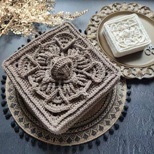 May include: Two crocheted squares, one brown and one white, with intricate patterns. The brown square is on a burlap mat with black pom-poms, and the white square is on a gold tray.