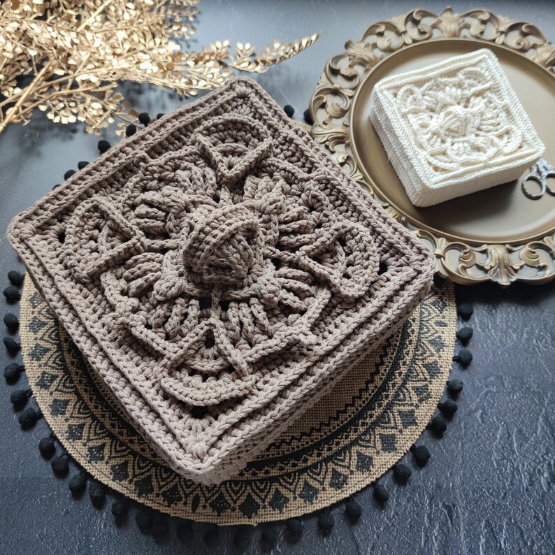 May include: Two crocheted squares, one brown and one white, with intricate patterns. The brown square is on a burlap mat with black pom-poms, and the white square is on a gold tray.