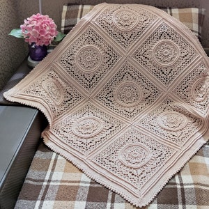 Pu&ograve; includere: Una coperta all'uncinetto di colore rosa chiaro con un delicato motivo a pizzo. La coperta &egrave; composta da motivi quadrati ripetuti con un design circolare al centro di ogni quadrato.