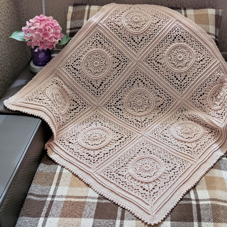 Pu&ograve; includere: Una coperta all'uncinetto di colore rosa chiaro con un delicato motivo a pizzo. La coperta &egrave; composta da motivi quadrati ripetuti con un design circolare al centro di ogni quadrato.
