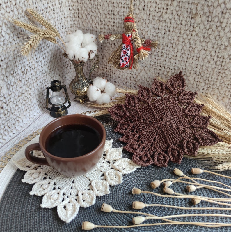 K&ouml;nnte beinhalten: Eine braune H&auml;keldeckchen mit einem wei&szlig;en H&auml;keldeckchen darauf. Eine Tasse Kaffee steht auf dem wei&szlig;en Deckchen. Die Deckchen liegen auf einer grauen Oberfl&auml;che mit getrockneten Blumenstielen.