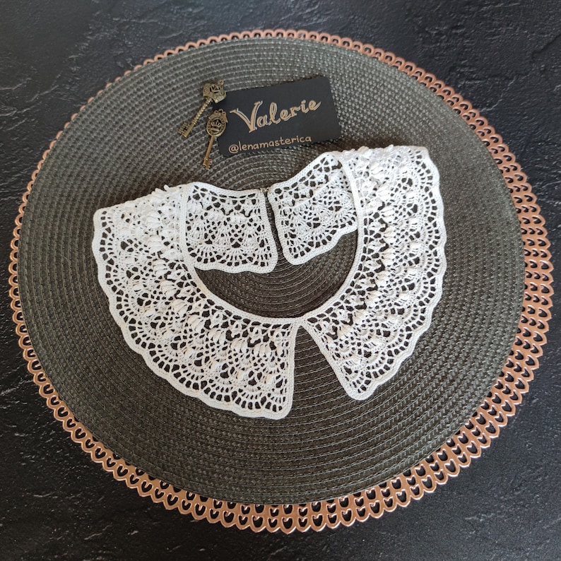 May include: White crocheted collar with a scalloped edge. The collar is on a green and brown woven placemat with a copper-colored trim. The text "Valerie @lenamasterica" is on a white card.