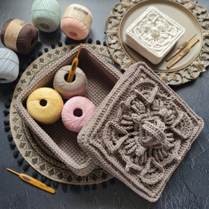 May include: A brown crocheted storage basket with a square crocheted lid. The basket is filled with yarn balls in various colors, including pink, white, and green. The basket is sitting on a black and white patterned surface.