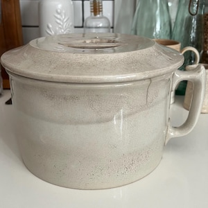 May include: A cream-colored ceramic soup tureen with a lid. The tureen has a handle and a slightly distressed, crackled finish. The background includes white tile and glass bottles.