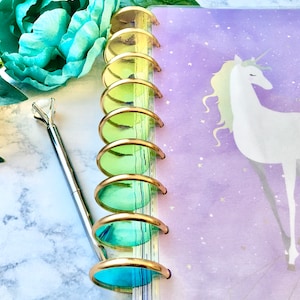 May include: A disc-bound planner featuring a purple cover with a white unicorn illustration and gold stars. The binding consists of a vertical row of gradient-colored discs transitioning from yellow to green to blue. A silver pen with a diamond topper.