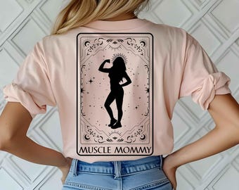 Może przedstawiać: Różowy T-shirt z czarną sylwetką kobiety uginającej biceps w ozdobnej ramie. Pod ramą nadrukowany jest tekst "MUSCLE MOMMY".