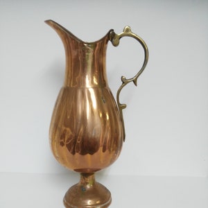 Op de afbeelding: Een koperkleurige kan met een decoratieve handgreep en een uitlopende tuit. De kan heeft een bolvormig lichaam en een voetstuk. De handgreep heeft een lichtere messingkleur. De kan is waarschijnlijk vintage.