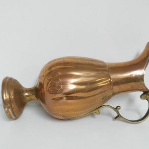 Op de afbeelding: Een koperkleurige kan met een bolvormige body, een uitlopende tuit en een decoratieve handgreep. De kan heeft een gepolijste afwerking, die licht reflecteert, en vertoont tekenen van ouderdom. De handgreep en voet lijken van messing te zijn. De kan is waarschijnlijk vintage.