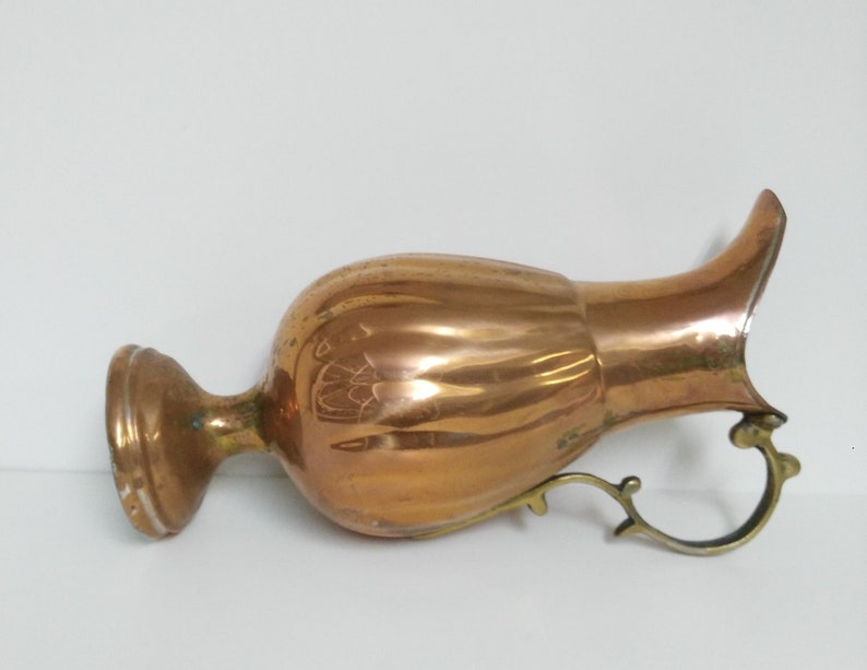 Op de afbeelding: Een koperkleurige kan met een bolvormige body, een uitlopende tuit en een decoratieve handgreep. De kan heeft een gepolijste afwerking, die licht reflecteert, en vertoont tekenen van ouderdom. De handgreep en voet lijken van messing te zijn. De kan is waarschijnlijk vintage.