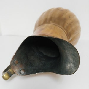 Op de afbeelding: Een vintage koperen kan met een afgeronde body en een brede tuit. De kan heeft een handvat en een aangetaste afwerking, die tekenen van ouderdom en gebruik vertoont. De binnenkant van de kan is donker, wat contrasteert met de warme tinten van de buitenkant.