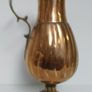 Op de afbeelding: Een koperkleurige kan met een decoratieve handgreep en een uitlopende tuit. De kan heeft een bolvormig lichaam en staat op een kleine, ronde voet. De handgreep is lichter van kleur, mogelijk messing. De kan is waarschijnlijk vintage of antiek.