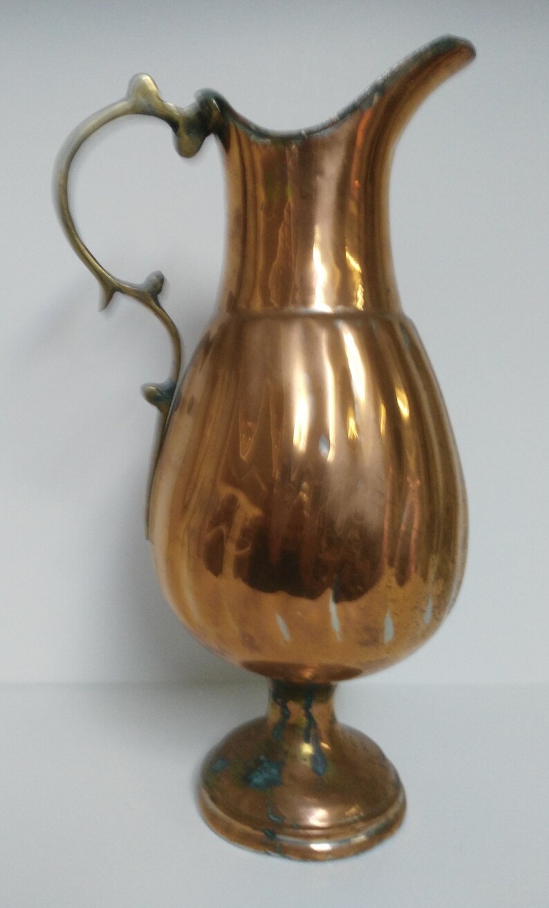 Op de afbeelding: Een koperkleurige kan met een decoratieve handgreep en een uitlopende tuit. De kan heeft een bolvormig lichaam en staat op een kleine, ronde voet. De handgreep is lichter van kleur, mogelijk messing. De kan is waarschijnlijk vintage of antiek.