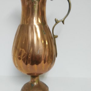 Op de afbeelding: Een koperkleurige kan met een bolvormige body en een slanke tuit. Het heeft een decoratieve messing handgreep en staat op een ronde voet. Het oppervlak van de kan reflecteert licht, wat de gepolijste afwerking benadrukt. Een vintage decoratief item.