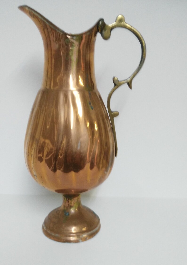 Op de afbeelding: Een koperkleurige kan met een bolvormige body en een slanke tuit. Het heeft een decoratieve messing handgreep en staat op een ronde voet. Het oppervlak van de kan reflecteert licht, wat de gepolijste afwerking benadrukt. Een vintage decoratief item.