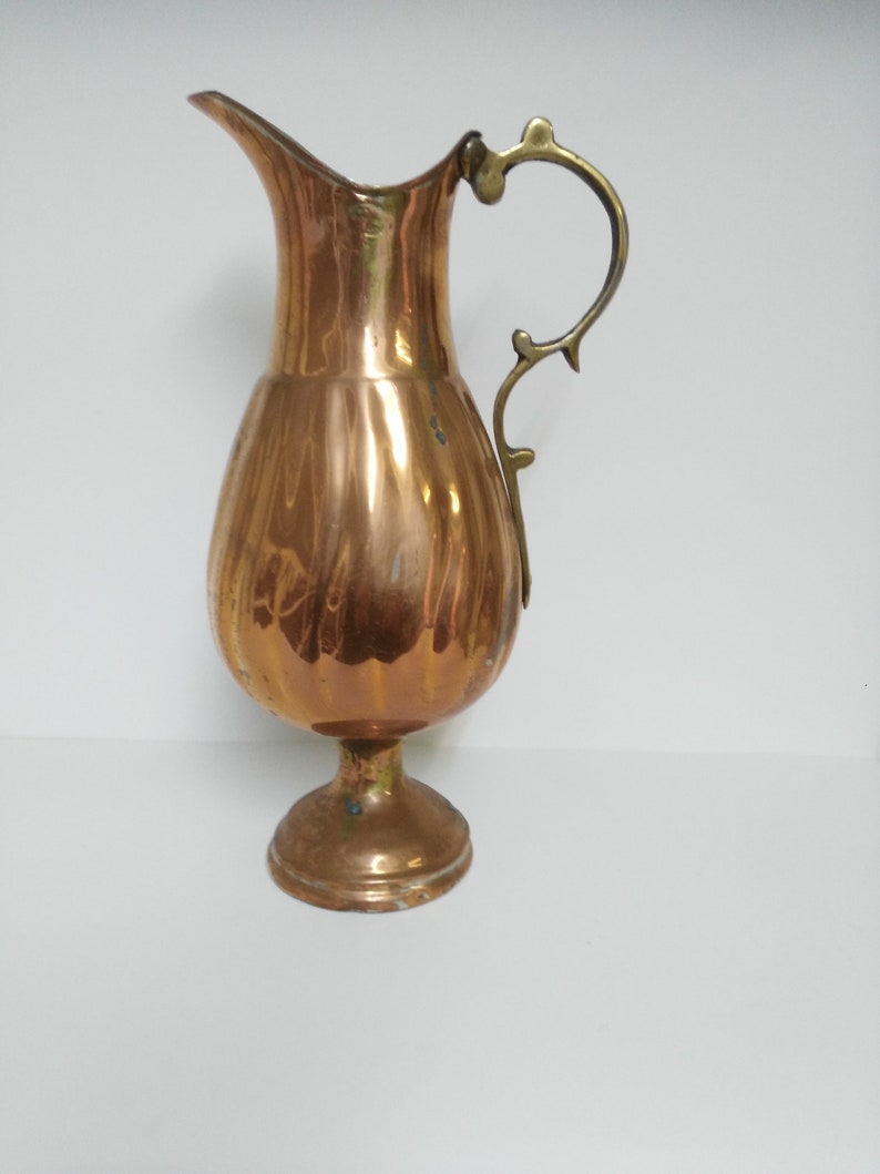 Op de afbeelding: Een koperkleurige kan met een decoratieve handgreep en een uitlopende tuit. De kan heeft een bolvormig lichaam en een voetstuk. De handgreep heeft een lichtere messingkleur. De kan is waarschijnlijk vintage.