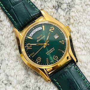 Op de afbeelding: Een goudkleurig polshorloge met een groene wijzerplaat en een groene leren band. De wijzerplaat toont de dag van de week en de datum. De wijzerplaat heeft de tekst "Pinaki" en "Automatic 25 Jewels".