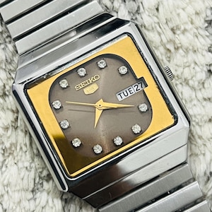 May include: A vintage Seiko 5 automatic watch with a silver stainless steel band. The watch face is square with a gold-colored border and a gray center, featuring diamond accents and the day and date display. The watch reads "TUE 27".