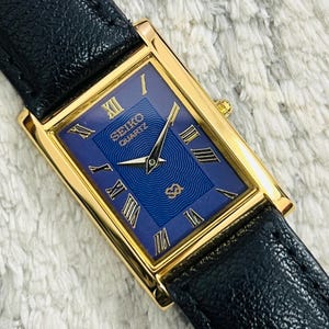 May include: A rectangular gold-toned Seiko Quartz watch with a blue face and black leather strap. The watch face features Roman numerals and the Seiko Quartz logo. The strap is black leather.