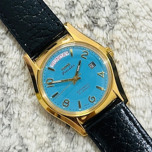 Op de afbeelding: Goudkleurig polshorloge met een lichtblauwe wijzerplaat en een zwarte leren band. De wijzerplaat toont "ZONDAG" en de datum "29". Het horloge heeft gouden wijzers en uurmarkeringen. Het horloge is gemarkeerd met "INDIA".