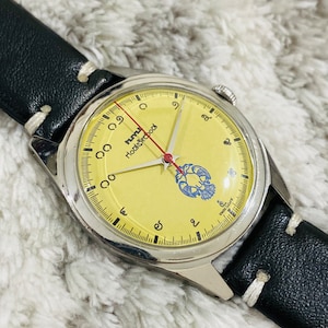 May include: A wristwatch with a yellow face and a black leather strap. The watch face has black markings and a red second hand. The watch face has the words "hurry" and "hokey pokey" printed on it.