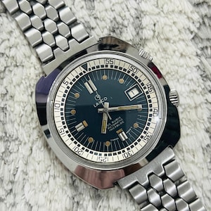 Puede incluir: Un reloj de pulsera plateado con una esfera negra y una correa de metal plateado. La esfera del reloj tiene marcas blancas y el texto "LANCO 25 RUBIS AUTOMATIC INCABLOC MADE T SWISS T".