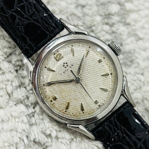 Puede incluir: Un reloj de pulsera plateado con una esfera blanca y una correa de cuero negra. El reloj tiene una esfera texturizada y marcadores de hora en números romanos.