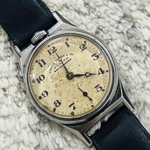 Könnte beinhalten: Eine silberne Armbanduhr mit einem schwarzen Lederarmband. Das Zifferblatt der Uhr ist weiß mit schwarzen römischen Ziffern als Stundenmarkierungen und schwarzen Zeigern. Das Zifferblatt der Uhr trägt außerdem den Text "Favre Leuba & Co.  Zenith Swiss Made".