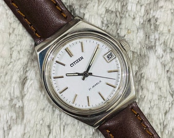 May include: A silver Citizen wristwatch with a white face and brown leather band. The watch face has black Roman numeral hour markers, black hands, and a date window at the 3 o'clock position. The watch face also says "Citizen" and "21 Jewels".