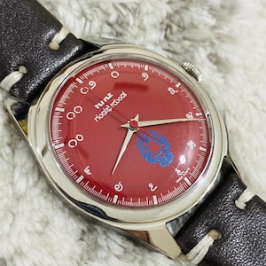 May include: A vintage wristwatch with a red face and silver casing. The watch has a dark brown leather strap with white stitching. The face includes a blue emblem and text in a foreign language.