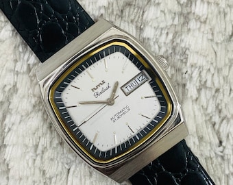 Puede incluir: Un reloj de pulsera plateado y dorado con una esfera blanca y una correa de cuero negro. La esfera del reloj tiene marcas negras y una ventana de fecha que dice "THU 12". La esfera del reloj también tiene el texto "RUTILE Kailash AUTOMATIC 21 JEWELS".