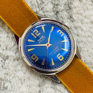 Op de afbeelding: Een polshorloge met een blauwe wijzerplaat, gouden accenten en een gele fluwelen band. De wijzerplaat van het horloge heeft de tekst "Kurth PILOT" en "PARA SHOCK 17 JEWELS".
