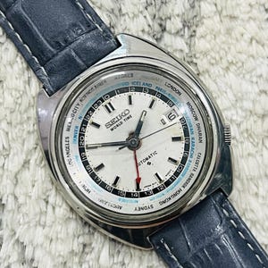May include: A silver Seiko World Time automatic watch with a white face and a black leather strap. The watch face features a 24-hour time zone ring with city names and a date window at the 3 o'clock position.