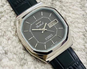 Puede incluir: Un reloj automático HMT Kailash plateado con esfera gris oscuro y correa de cuero negro. La esfera muestra el día y la fecha, con el texto "TUE 25". El reloj tiene forma rectangular.
