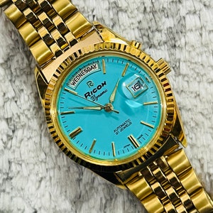 May include: A gold-tone wristwatch with a blue face. The watch has a day and date window, and the text "Ricoh Dynamic Automatic 21 Jewels Japan Made" is printed on the face.