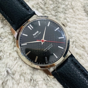 May include: A black-faced wristwatch with a silver case and black leather strap. The watch face has white markings and the text "HMT SLIM PARA SHOCK 17 JEWELS".