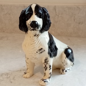 Puede incluir: Figura de cerámica de un perro sentado, principalmente blanco con manchas negras. La cabeza y las orejas son negras. El acabado brillante sugiere un objeto decorativo, ideal para una estantería.