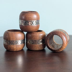 May include: Set of four wooden napkin rings with a silver band. The rings are brown and have a decorative design on the silver band.
