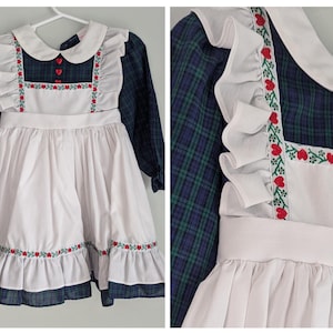 May include: A vintage-style dress with a white bib-style apron and a navy blue plaid bodice. The apron is adorned with red floral embroidery and a ruffled trim. The dress has long sleeves and a white collar, with red heart-shaped buttons.