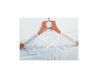 Può includere: Colletto in pizzo bianco con fiori ricamati e dettagli di perle. Il colletto è su un appendiabiti in legno.