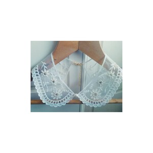 May include: White lace collar with pearl and floral embellishments. The collar is on a wooden hanger.