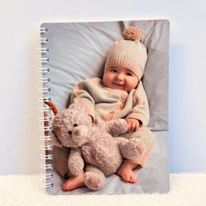 Peut inclure: Un bébé portant un bonnet et un pull en tricot rose et blanc tient un ours en peluche. Le bébé est allongé sur une couverture bleu clair.