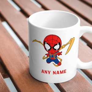 Puede incluir: Taza de cer&aacute;mica blanca con una imagen de dibujos animados de Spider-Man con un traje rojo y azul con detalles dorados. La taza tiene un asa y el texto "ANY NAME" impreso en la parte inferior.
