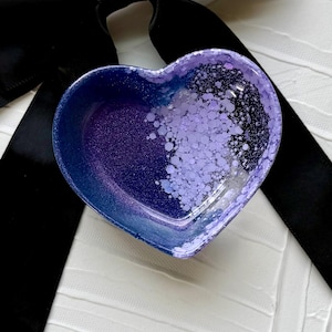 Pode incluir: Uma tigela em forma de coração com um gradiente de azul escuro a roxo, salpicada com glitter roxo claro. A tigela está sobre uma superfície branca texturizada e uma fita preta.