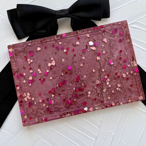May include: A rectangular tray with a mauve base, embedded with heart-shaped glitter in shades of pink and gold. A black bow and ribbon are in the background, creating a contrast with the tray's design.