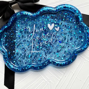 May include: A cloud-shaped tray with a glittery blue surface. The tray has the words "You are Loved" in white script, along with two white heart shapes. A black ribbon is visible in the background.