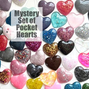 May include: A collection of heart-shaped pocket stones in various colors, including pink, blue, and gold, with glitter and sparkle effects. The image features the text "Mystery Set of Pocket Hearts."