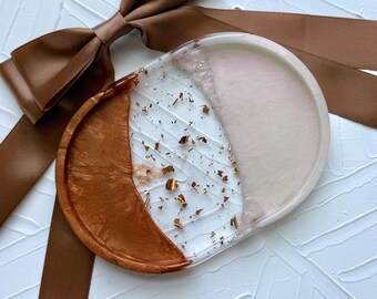 May include: A decorative resin tray with a unique design. The tray is divided into three sections: a copper-colored section, a clear section with gold flakes, and a blush-colored section. A brown satin ribbon and bow are in the background.