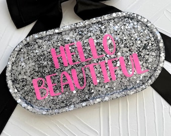May include: A glittery, oval-shaped tray with the words "HELLO BEAUTIFUL" in bright pink lettering. The tray is filled with silver glitter and has a dark border. A black bow and ribbon are in the background.