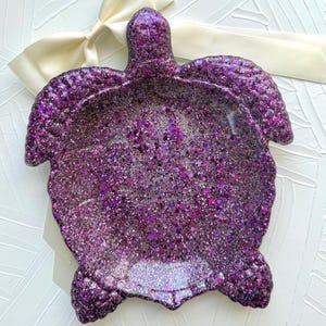 May include: A purple glitter turtle-shaped tray with a raised head and flipper-like sides. The tray is covered in a mix of purple glitter and sits on a white surface with a cream-colored ribbon.