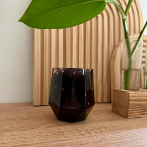 May include: A small, faceted, dark black vessel with a glossy finish and embedded copper-colored metallic flakes. The container has a geometric, multi-sided shape with a wide opening at the top and a narrower base, featuring a smooth, reflective surface.